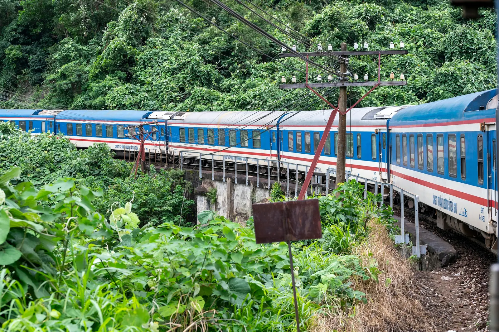 A blue and white passenger train crossing a forested railway bridge near Lang Co.